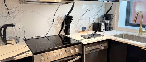 A modern kitchen with a built-in stove, oven, and dishwasher, a marble backsplash, and a window with blinds.