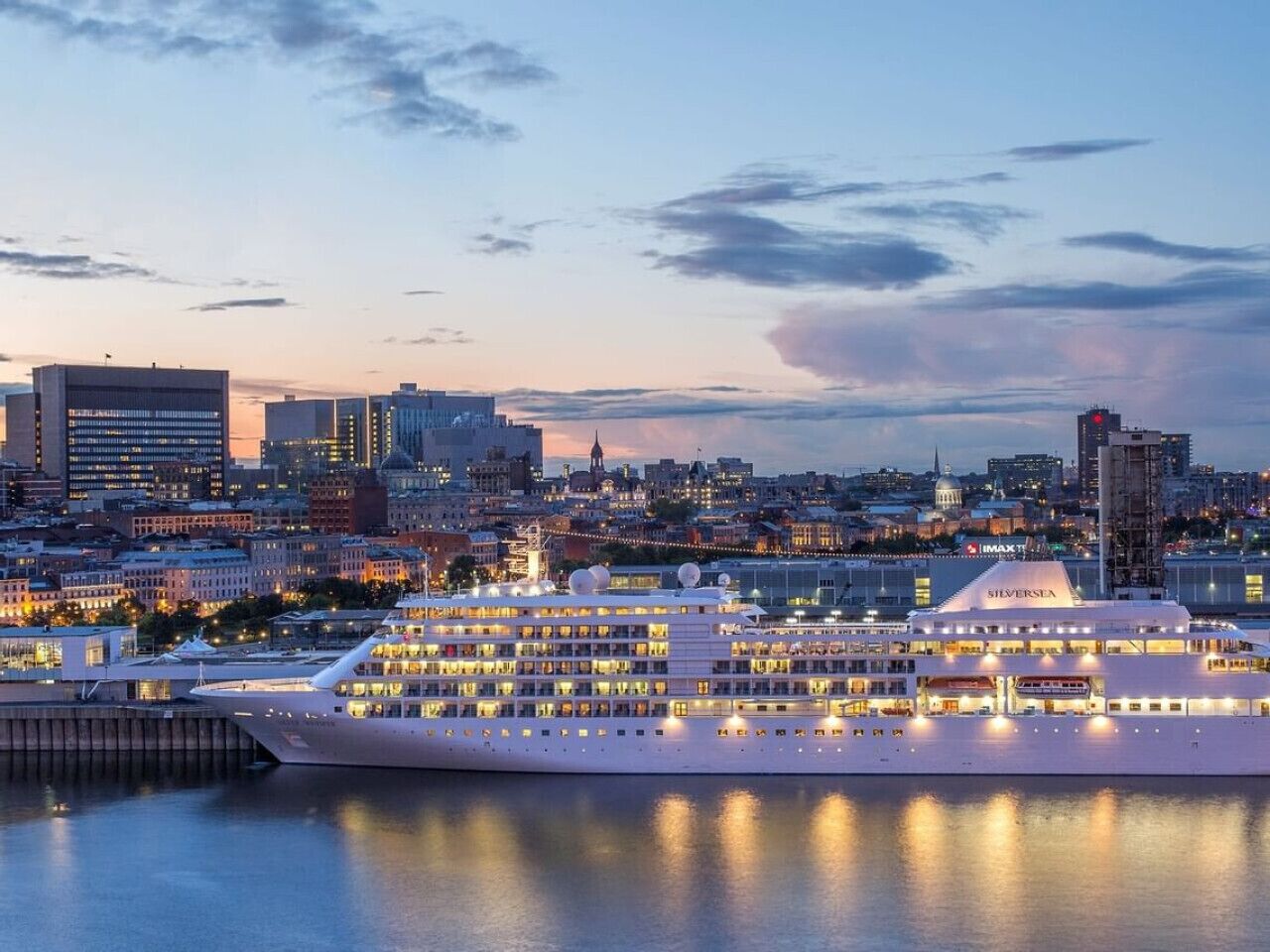 A large cruise ship docked at a port with a city skyline in the background.