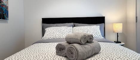 A neatly made bed with rolled towels, a patterned bedspread, and a bedside lamp.