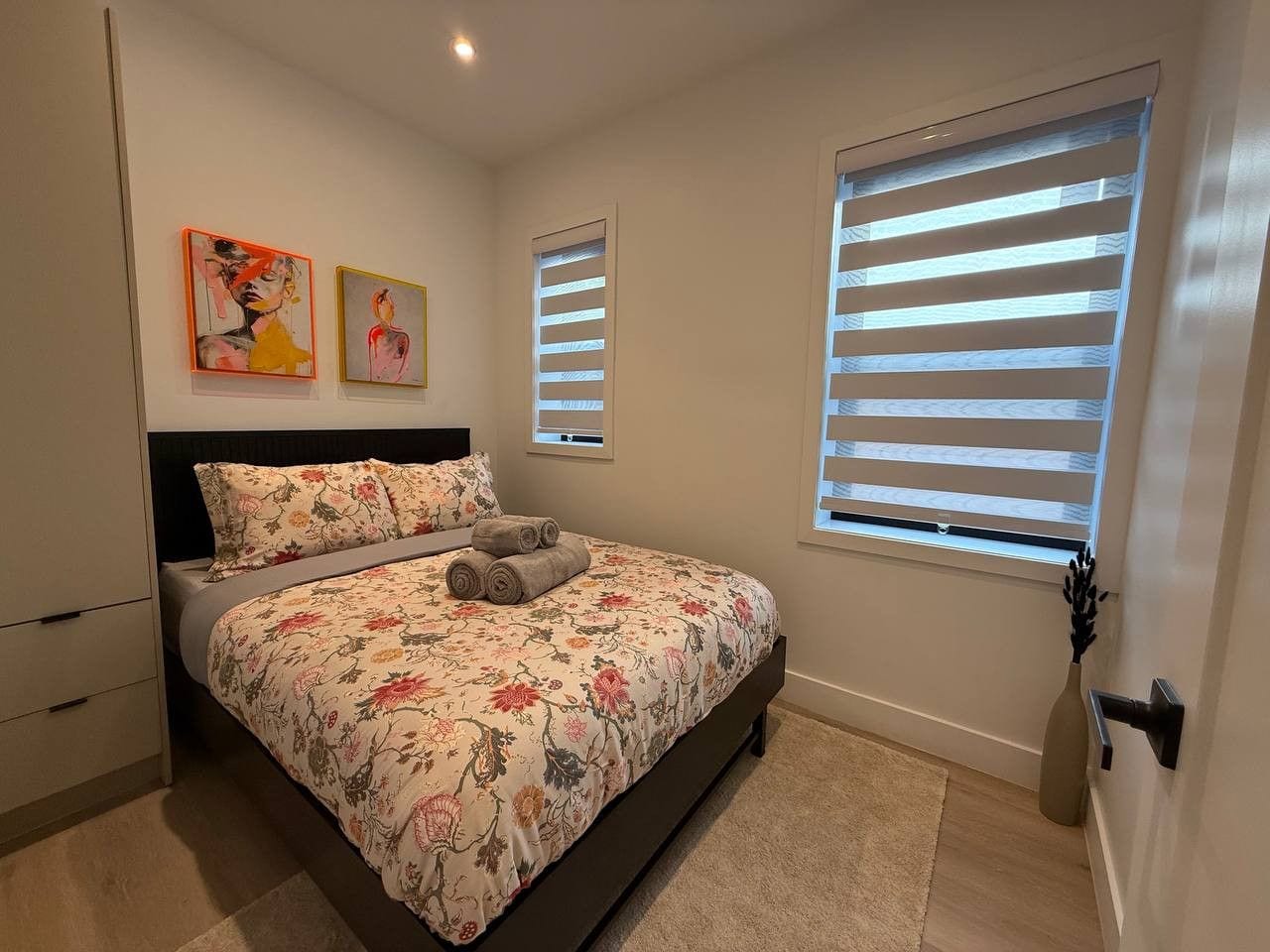 Un dormitorio con cama, mesita de noche, cómoda y dos ventanas con persianas.