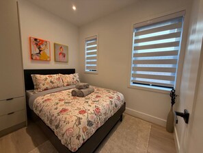 Une chambre à coucher comprenant un lit, une table de chevet, une commode et deux fenêtres équipées de stores.