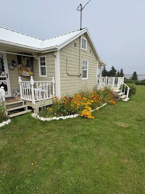 A single-story house with a porch, a small garden with colorful flowers, and a view of the water. - Oceanview Cottage - Beach - Peaceful - Amazing Views (Malpeque)