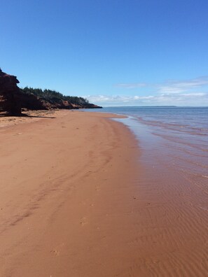 En bred sandstrand med milda vågor, en skogbeklädd klippa och en klarblå himmel.
