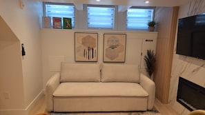 Ein Wohnzimmer mit einem beigefarbenen Sofa, zwei abstrakten Gemälden, einem Kamin und einem an der Wand montierten Fernseher.