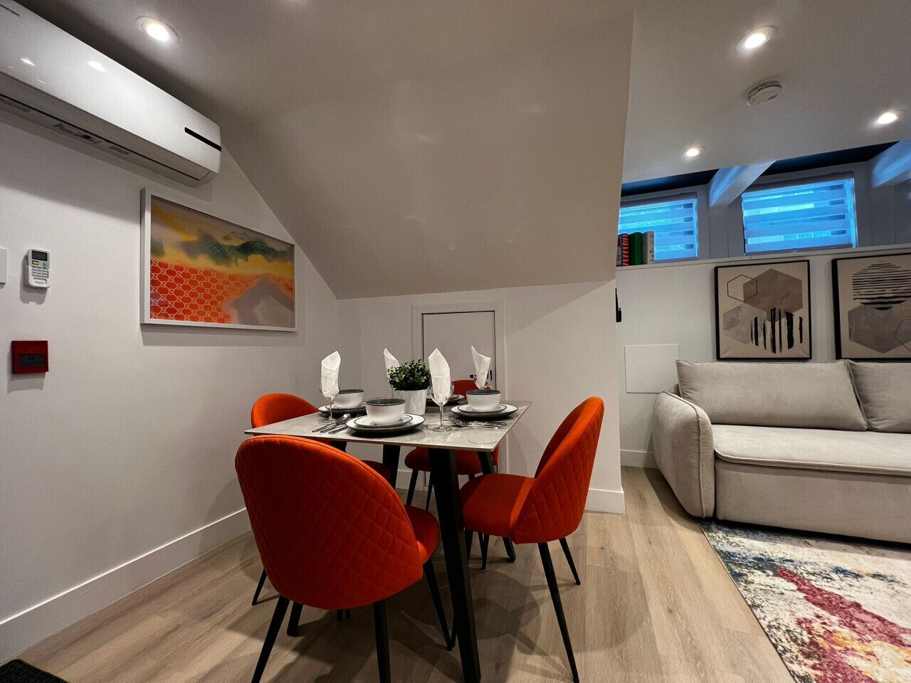 A dining area with a table set for four, surrounded by orange chairs, a sofa, and a patterned rug.
