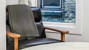 Une pièce avec une fenêtre, une chaise recouverte d’une couverture, un lit et un tableau encadré au mur.