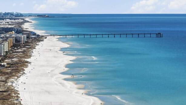 Una playa con un muelle que se adentra en el agua, edificios a lo largo de la orilla y un cielo despejado.