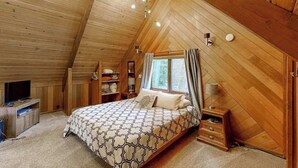 Un dormitorio con techo inclinado, una cama con colcha estampada, una mesita de noche con lámpara y un televisor sobre un mueble.
