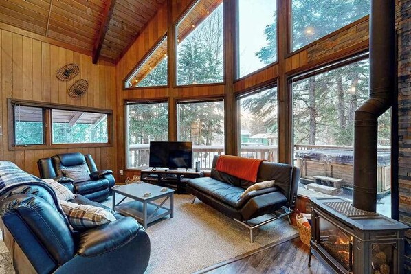 Ein gemütliches Wohnzimmer mit Kamin, Ledersofas, einem Flachbildfernseher und großen Fenstern mit Blick auf den Wald.
