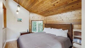 Una camera da letto con soffitto in legno, un letto con biancheria bianca, un comodino e una finestra con vista sul verde.