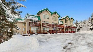 A multi-story hotel with balconies, a snow-covered driveway, and surrounding trees. - First Tracks Chalet | Renovated Base Condo (Girdwood)