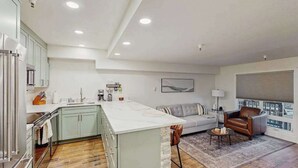 A modern kitchen with a central island, a dining area with a sofa and a coffee table, and a view through a window. - First Tracks Chalet | Renovated Base Condo (Girdwood)
