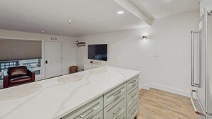 A modern kitchen with a large island, a flat-screen TV, and a sliding door leading to a balcony. - First Tracks Chalet | Renovated Base Condo (Girdwood)