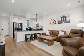 Una sala moderna con un sofá de cuero marrón, una mesa de centro de madera y una cocina con armarios blancos y electrodomésticos de acero inoxidable.