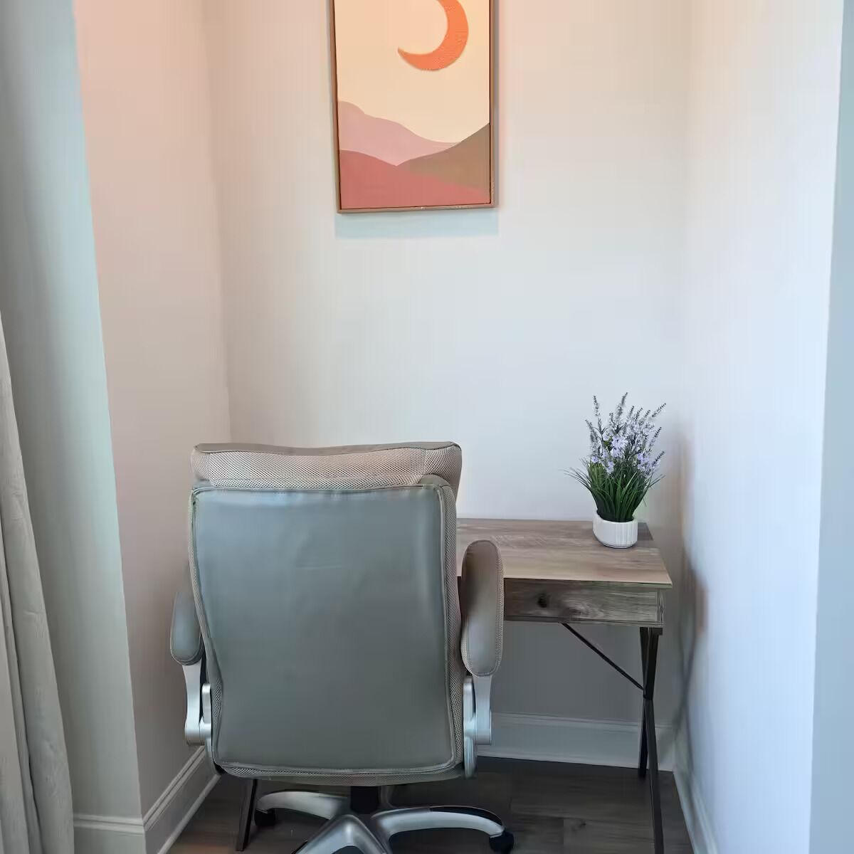 A corner office with a grey office chair, a wooden desk, and a wall-mounted painting.
