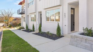 Un edificio residencial de varios pisos con fachada blanca, una entrada visible y un pequeño jardín con arbustos podados.