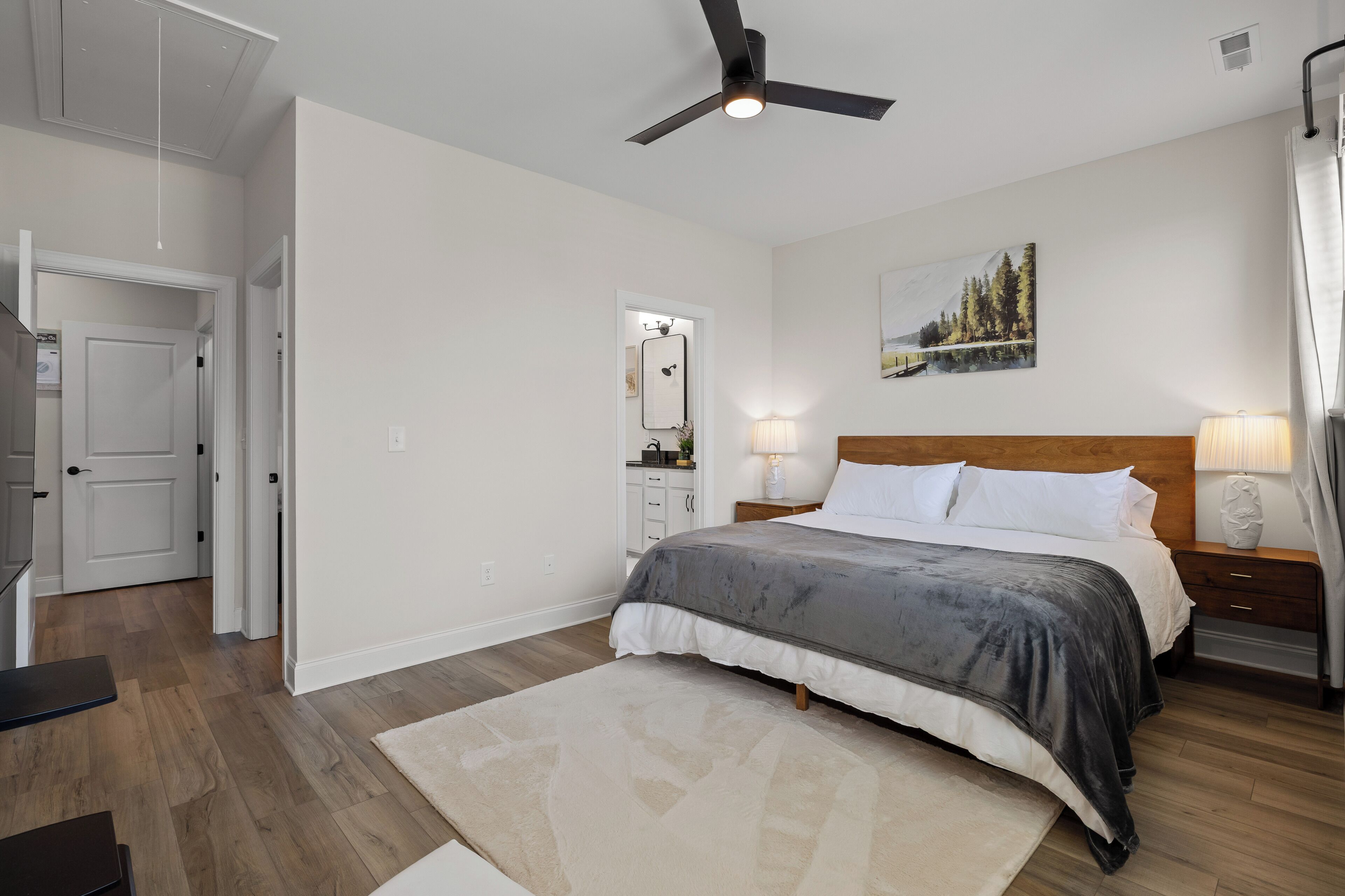 A bedroom with a bed, bedside lamps, a ceiling fan, a painting, and a wooden floor.