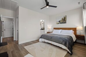 A bedroom with a bed, bedside lamps, a ceiling fan, a painting, and a wooden floor.