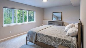 A bedroom with a large window, a bed with patterned bedding, a dresser, and a mirror. - Penguin Ridge Estate | Full 4BR Duplex Compound (Girdwood)