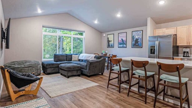 A modern kitchen with wooden cabinets, a stainless steel refrigerator, and a dining area with bar stools. - Penguin Ridge Estate | Full 4BR Duplex Compound (Girdwood)