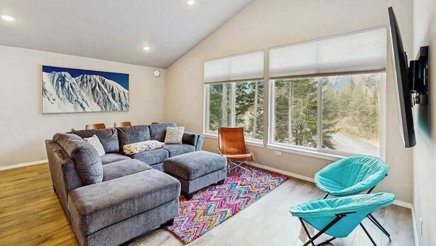 Un salon moderne avec un canapé d’angle gris, un tapis coloré, un tableau représentant une montagne et une grande fenêtre donnant sur les arbres.