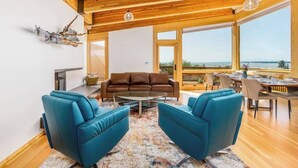 Ett modernt vardagsrum med ett träpanelat tak, en brun lädersoffa, två blå fåtöljer, ett soffbord och en matplats med utsikt över havet.
