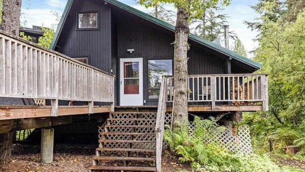 A cabin with a wooden deck, a front door, and a staircase leading to the entrance. - Alpine Meadows A-Frame | Walk to Slopes (Girdwood)