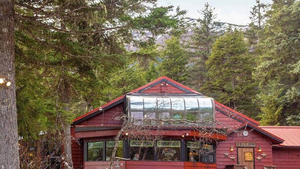 A red wooden building with a glass roof, a 'Welcome' sign, and a view of the forest. - North 413 | True Ski-In/Out | Gas Fireplace (Girdwood)