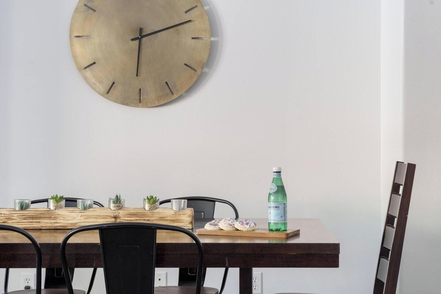 A dining area with a wooden table, chairs, a large wall clock, and a bottle of water.