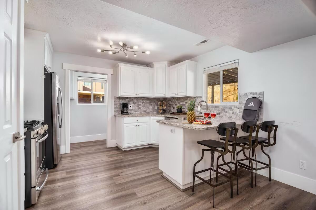 A modern kitchen with white cabinets, a central island with bar stools, a built-in stove, and a window overlooking the backyard.