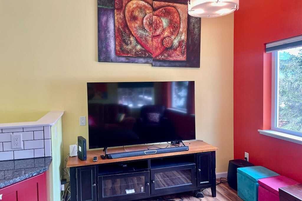 Een woonkamer met een flatscreen televisie op een houten standaard, een rode muur, een raam en een schilderij boven de televisie.