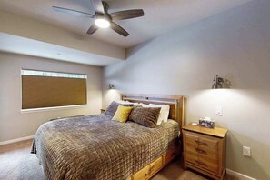 Une chambre à coucher comprenant un lit, une table de chevet, un ventilateur de plafond, une fenêtre avec des stores et un luminaire fixé au mur.