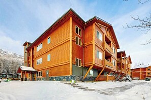 A multi-story wooden lodge with a balcony on the second floor, a covered entrance, and a snow-covered ground. - Hanging Glacier Chalet - Group Lodge - Sauna (Girdwood)