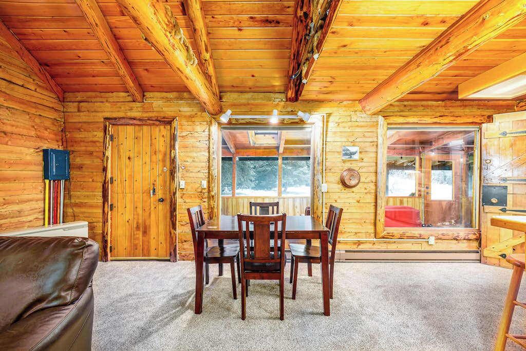 A wooden cabin interior with a dining table, chairs, a couch, and a fireplace.