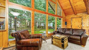 A log cabin interior with a leather sofa, a coffee table, and large windows offering a view of the forest.