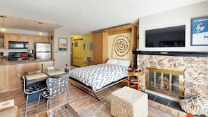 Ein kompaktes Hotelzimmer mit Küchenzeile, einem Bett mit gemusterten Betttextilien, einem Kamin und einem Wandfernseher.