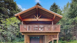 Una cabaña de madera de dos pisos con un balcón, rodeada de un bosque frondoso.