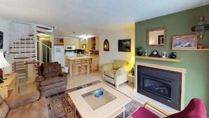 A living room with a fireplace, a sofa, a coffee table, and a kitchen in the background.