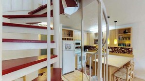 A kitchen with a central island, a staircase with red accents, and a dining area with wooden chairs.