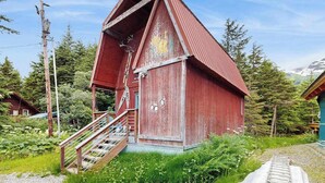 A red wooden building with a steep roof, a staircase leading to a porch, and a small window. - Kabina on Vail | Lofted Cottage | Spiral Stairs (Girdwood)