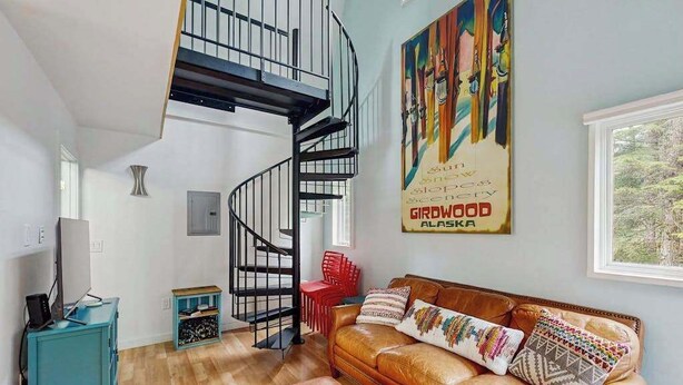 A living room with a spiral staircase, a sofa with patterned cushions, a small table, and a painting on the wall. - Kabina on Vail | Lofted Cottage | Spiral Stairs (Girdwood)
