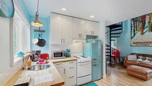 A modern kitchen with white cabinets, a stove, and a microwave. There is a dining area with a red chair and a couch. - Kabina on Vail | Lofted Cottage | Spiral Stairs (Girdwood)