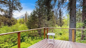 A wooden deck with a railing, overlooking a forested area with tall trees and a mountain in the background.