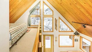 A high-ceilinged room with a sloped wooden roof, a bed, a window with a view of snow-covered trees, and a door.