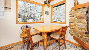 A dining area with a wooden table and chairs, large windows with snowy views, a stone fireplace, and a wooden ceiling.