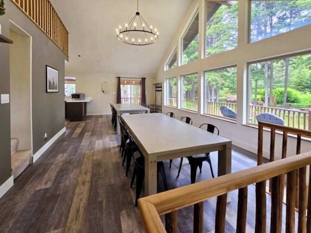 A spacious room with a long table, chairs, a chandelier, large windows, and a wooden railing.
