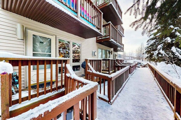 A multi-story building with a snow-covered deck and railings, a visible window, and a snowy landscape. - North 409 | Ski-In/Out | Vintage Ski Decor (Anchorage)