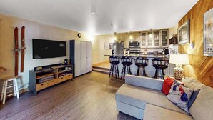 A modern living room with a flat-screen TV, a sofa, and a dining area with bar stools. - North 409 | Ski-In/Out | Vintage Ski Decor (Anchorage)