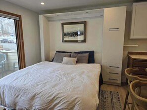 A bedroom with a bed, a desk, and a chair. There is a window with a view of the outside.