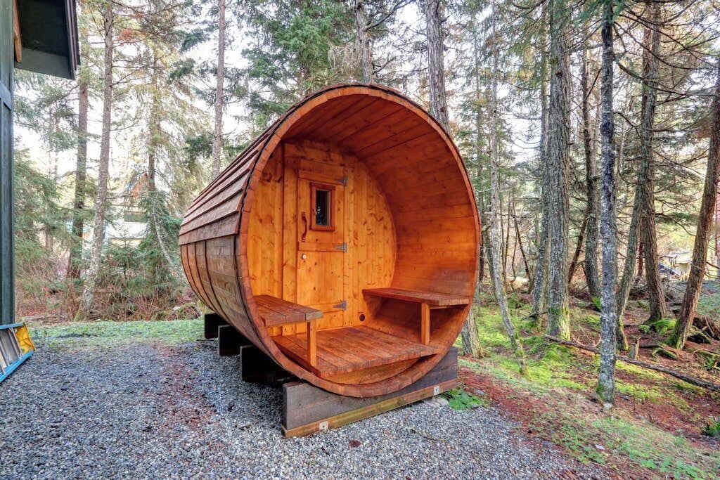 Une structure en forme de tonneau, faite de bois, avec une porte, des fenêtres et des sièges à l’intérieur, située au cœur d’une forêt.